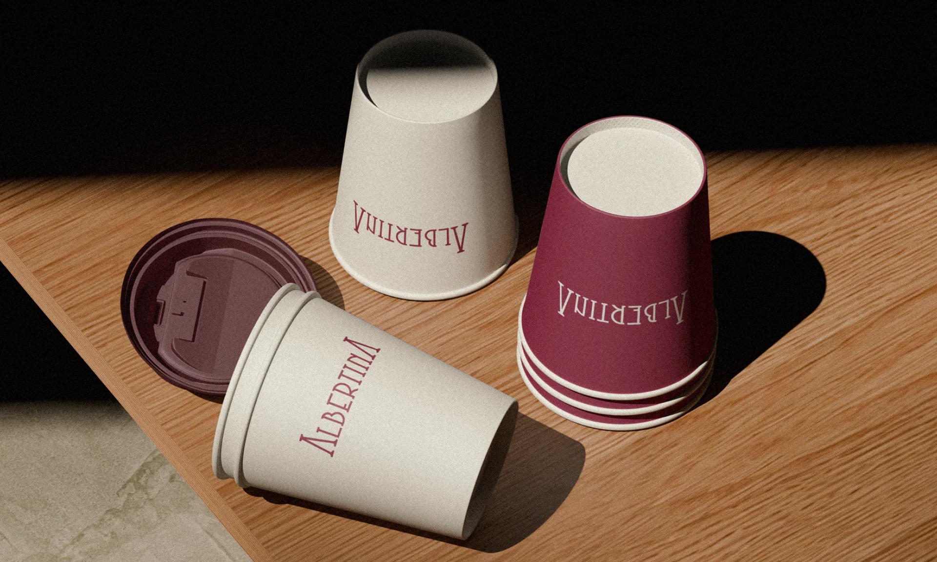 Takeaway cups in cream and burgundy with the wordmark, on a wooden table in hard sunlight.